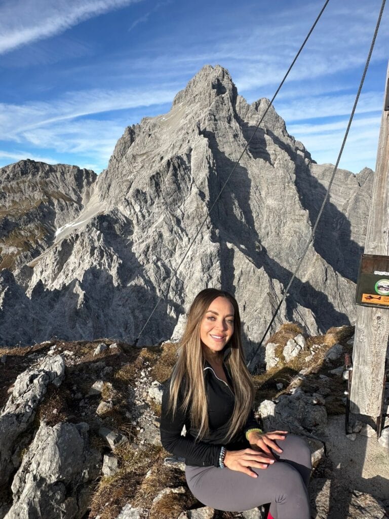 Eine lächelnde Frau mit langen Haaren sitzt vor einem beeindruckenden Berg in der Sonne.